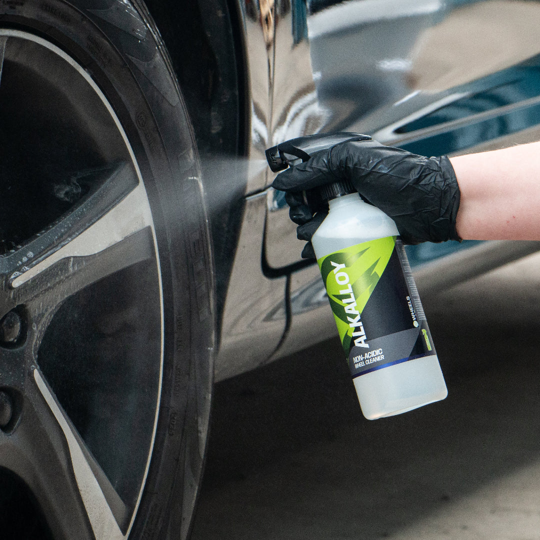 A person wearing a black glove sprays a cleaning solution from a white spray bottle labeled 'Alkalloy' onto a car wheel in a garage setting.