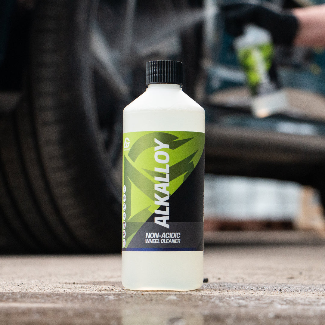 A bottle of Alkalloy Non-Acidic Wheel Cleaner stands on wet pavement in front of a blurred car and a person spraying water, demonstrating its use.