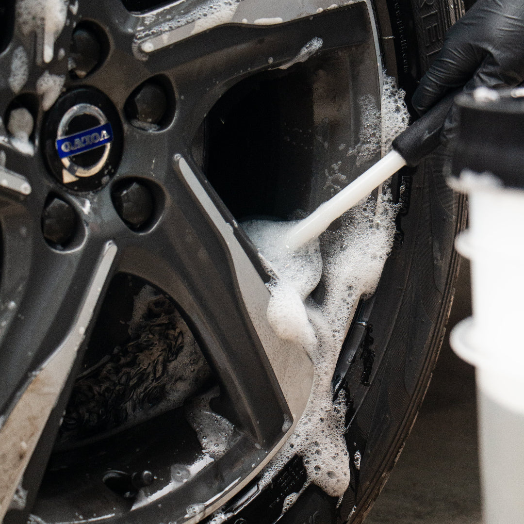 A close-up of a car wheel being cleaned with a foam cannon covered in white soap foam, with a black gloved hand holding the brush. The Volvo logo is visible on the wheel hub.