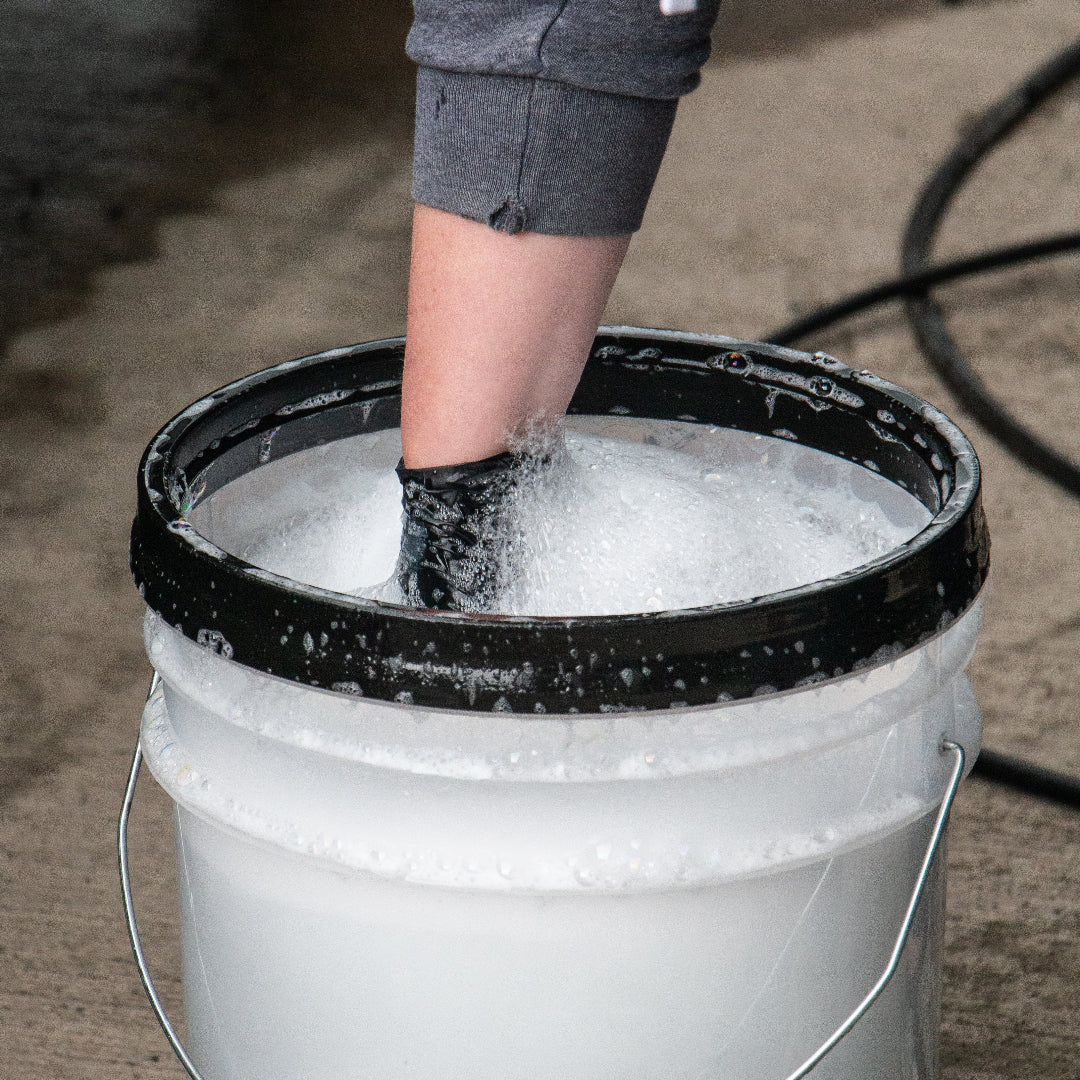 A person's arm with a black glove dipping into a white bucket filled with soapy water on a concrete floor.