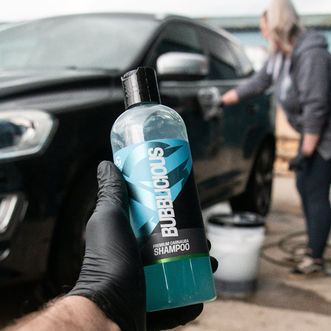 A hand wearing a black glove holds a bottle of Bubblicious Car Shampoo in the foreground, with a black car and a person washing it in a real-world outdoor setting in the background.