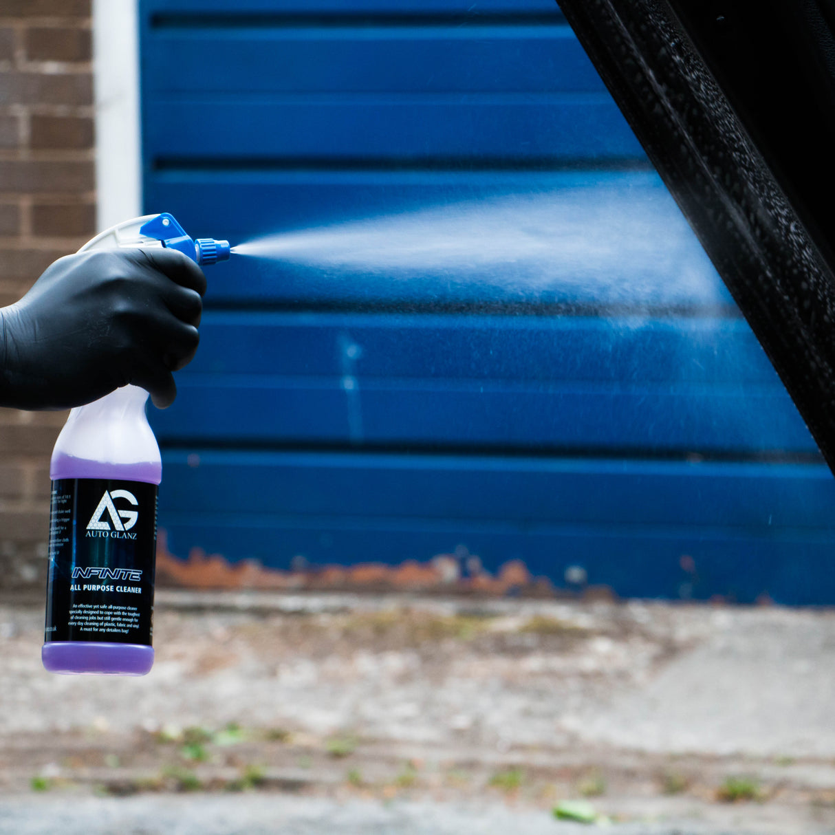 A hand wearing a black glove holds a spray bottle and sprays a fine mist onto a vehicle door, with a blue garage door and brick wall in the background.