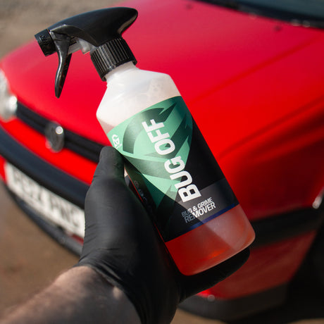 Hand holding a 'BUG OFF' spray bottle in front of a red car