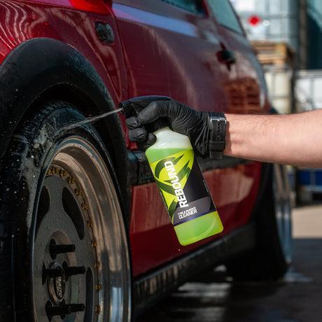 A person wearing a black glove sprays a green cleaning solution from a bottle onto a red car wheel in an outdoor setting.