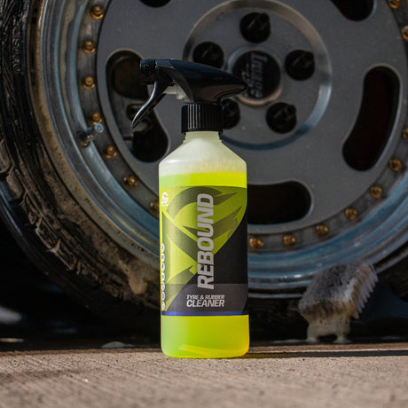 A bottle of Rebound Cleaner with a black spray top and neon yellow-green liquid, standing in front of a car wheel on a concrete surface.