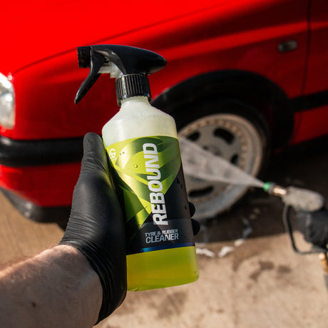 A hand wearing a black glove holds a bottle of REBOUND Car Cleaner in the foreground, with a red car and a water spray hose blurred in the background.