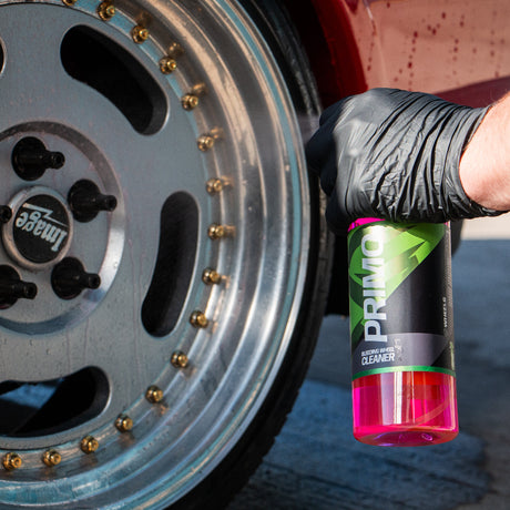 A close-up of a hand wearing a black glove holding a bottle of Primo car wheel cleaner spray, positioned next to a custom silver wheel with gold-colored lug nuts.