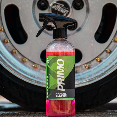 A bottle of Primo Car Wheel Cleaner with pink liquid and a green label, placed in front of a blurred car wheel.