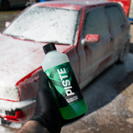 A hand wearing a black glove holds a bottle of Piste snow foam in front of a red car covered in white snow foam in an outdoor setting.