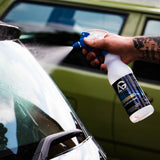 A tattooed hand holding a white spray bottle with a blue trigger sprayer, spraying a glass cleaner onto a windshield. The background shows a blurred olive green vehicle.