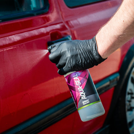 A hand wearing a black glove sprays a clear bottle of Aqua Seal car sealant onto the side of a red car.