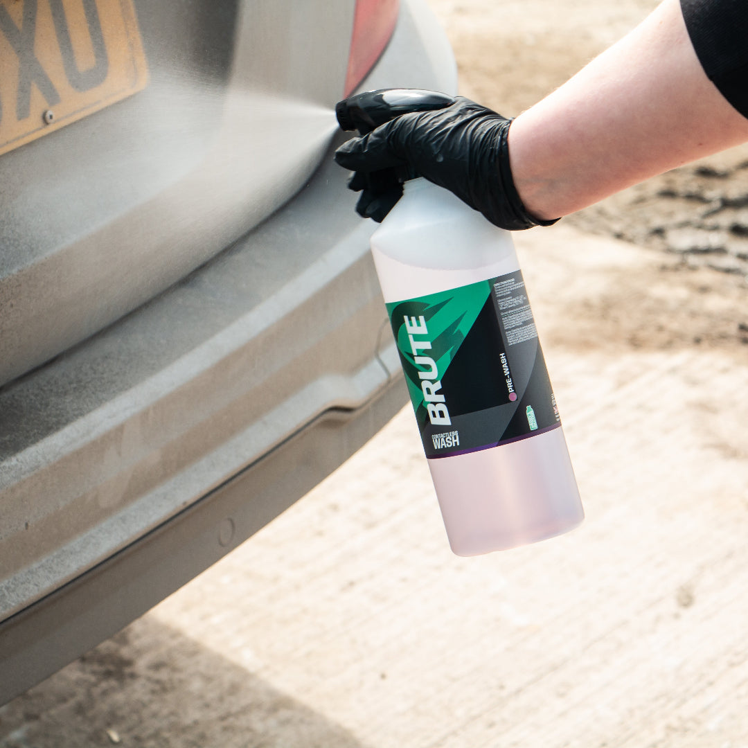 A person wearing a black glove sprays a car bumper cleaner from a white bottle labeled BRUTE onto a grey vehicle bumper in an outdoor setting.