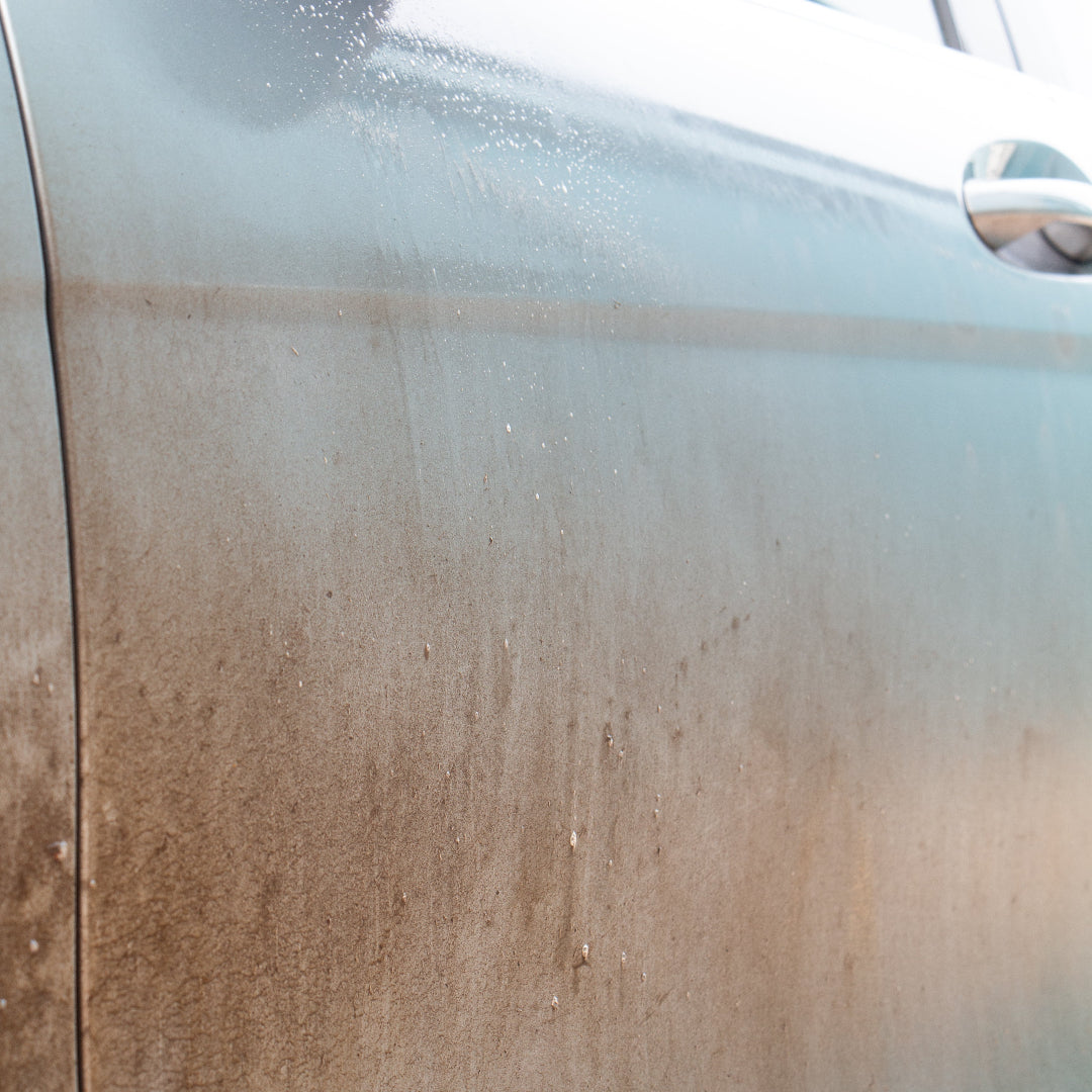 Close-up of a dirty metallic grey car door panel covered in dust and grime, with a chrome door handle visible on the right.