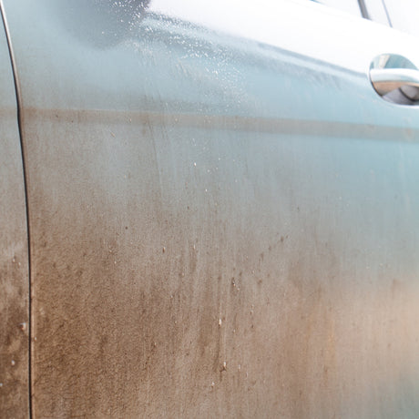 Close-up of a dirty metallic grey car door panel covered in dust and grime, with a chrome door handle visible on the right.