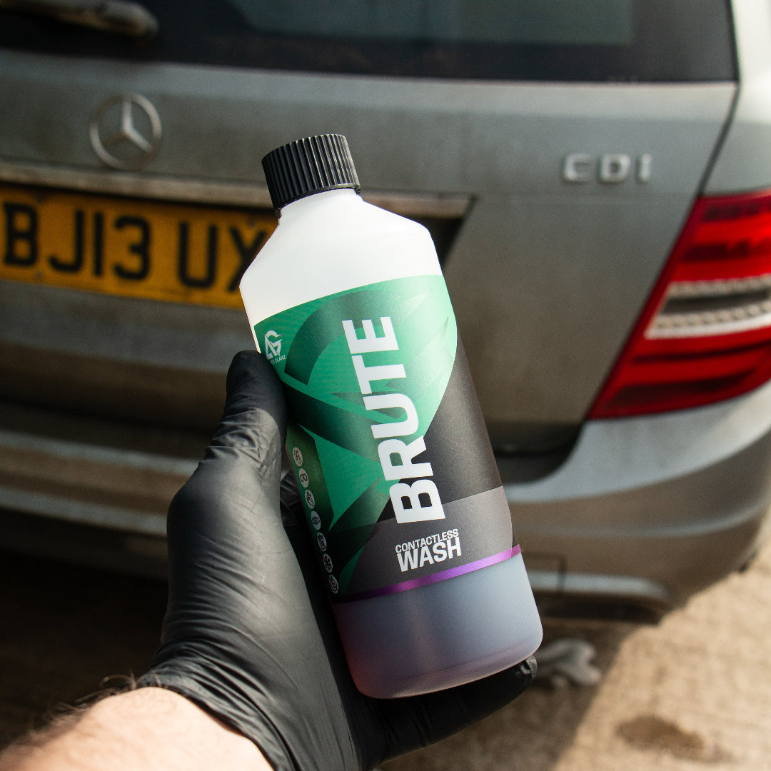 A hand wearing a black glove holds a bottle of BRUTE car wash in front of a silver Mercedes-Benz car, with the rear license plate and taillight visible in the background.