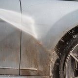 A close-up of a silver car door and wheel area being washed with a strong water spray, showing dirty water streaks running down the door panel and wheel well.