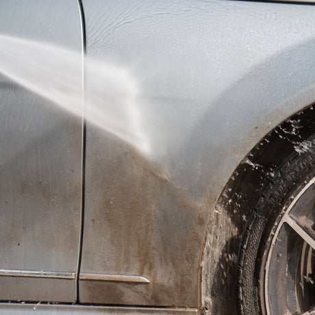 A close-up of a silver car door and wheel area being washed with a strong water spray, showing dirty water streaks running down the door panel and wheel well.