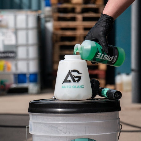 A person wearing a black glove pours green snow foam into a white Auto Glanz foam cannon mounted on a white washing bucket with a black lid.