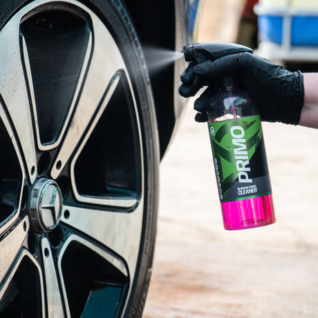 A hand wearing a black glove holds a bottle of Primo Car Care Wheel Cleaner spray next to a car wheel, with the pink and green cleaner liquid visible in the bottle.