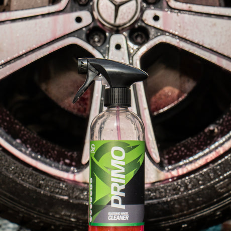 A bottle of PRIMO Wheel Cleaner spray with a black trigger sprayer and green/black label, standing in front of a wet car wheel with Mercedes-Benz branding.