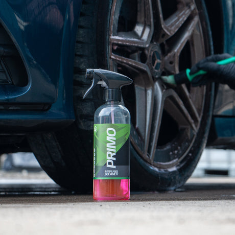 A bottle of Primo Wheel Cleaner spray stands on the ground next to a car wheel, with a person wearing black gloves using a green applicator on the tire. The bottle has a black spray top, a clear body showing pink liquid at the bottom, and a green and black label with the brand name 'PRIMO' in white.