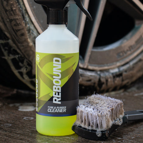 A bottle of REBOUND Car Tyre Cleaner with a black spray top and a white-to-yellow liquid, shown next to a dirty cleaning brush in front of a car wheel on wet pavement.