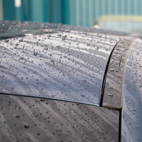 Close-up of the rear roof and window area of a dark blue car covered in water beads, with a teal wall and part of a red vehicle blurred in the background.
