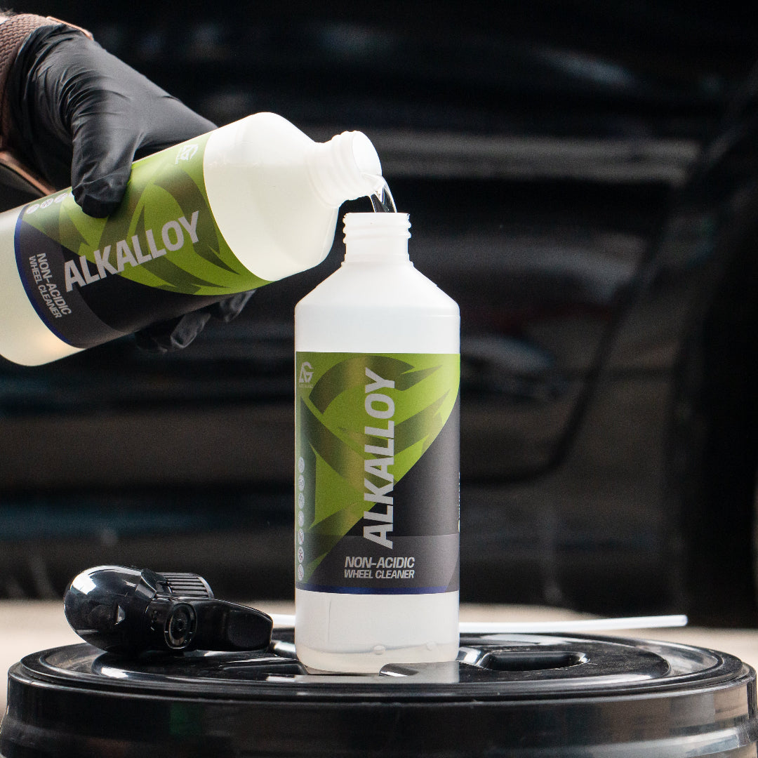 A person wearing a black glove pours liquid from a bottle labeled ALKALLOY into a larger white bottle on a black barrel, with a blurred car in the background.