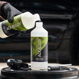 A person wearing a black glove pours liquid from a bottle labeled ALKALLOY into a larger white bottle on a black barrel, with a blurred car in the background.