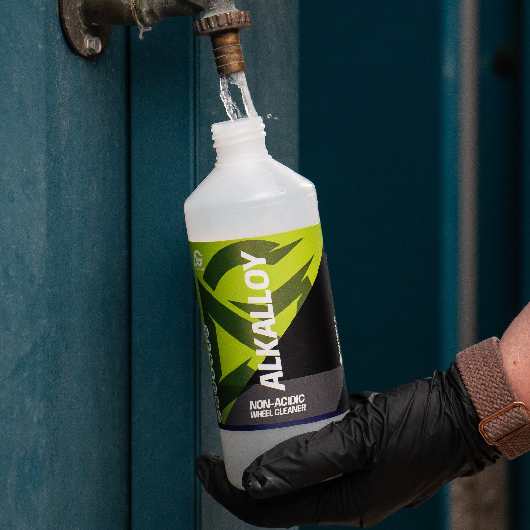 A person wearing black gloves holds a bottle of ALKALLOY Non-Acidic Wheel Cleaner under a metal faucet, with water flowing into the bottle, set against a dark teal wooden door.