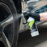 A person wearing a black glove sprays a cleaning solution from a white spray bottle labeled 'Alkalloy' onto a car wheel in a garage setting.