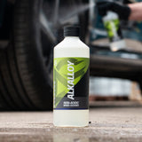 A bottle of Alkalloy Non-Acidic Wheel Cleaner stands on wet pavement in front of a blurred car and a person spraying water, demonstrating its use.