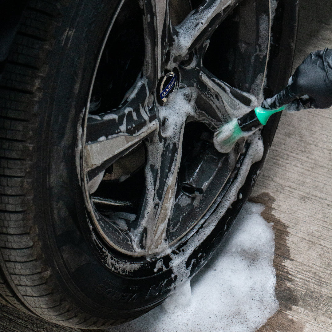 A close-up of a car wheel being washed with foam and a green-branded brush, with wet concrete and foam pooling below.
