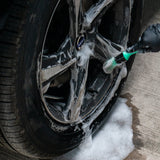 A close-up of a car wheel being washed with foam and a green-branded brush, with wet concrete and foam pooling below.