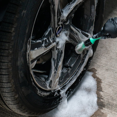 A close-up of a car wheel being washed with foam and a green-branded brush, with wet concrete and foam pooling below.