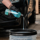 A person wearing a black glove pours blue liquid from a bottle labeled Bubblicious into a bucket, with a dark car in the background.