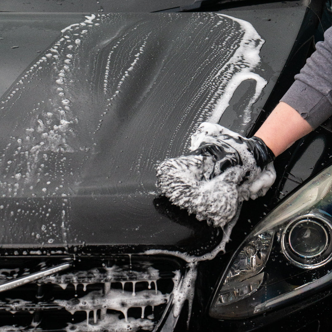 A close-up of a black car being washed, with white soap foam covering the bonnet and grill. A person's gloved hand is washing the front, and part of the car headlight is visible.