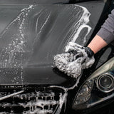 A close-up of a black car being washed, with white soap foam covering the bonnet and grill. A person's gloved hand is washing the front, and part of the car headlight is visible.