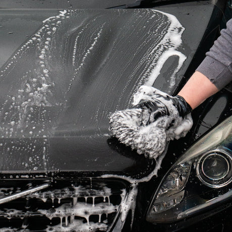 A close-up of a black car being washed, with white soap foam covering the bonnet and grill. A person's gloved hand is washing the front, and part of the car headlight is visible.