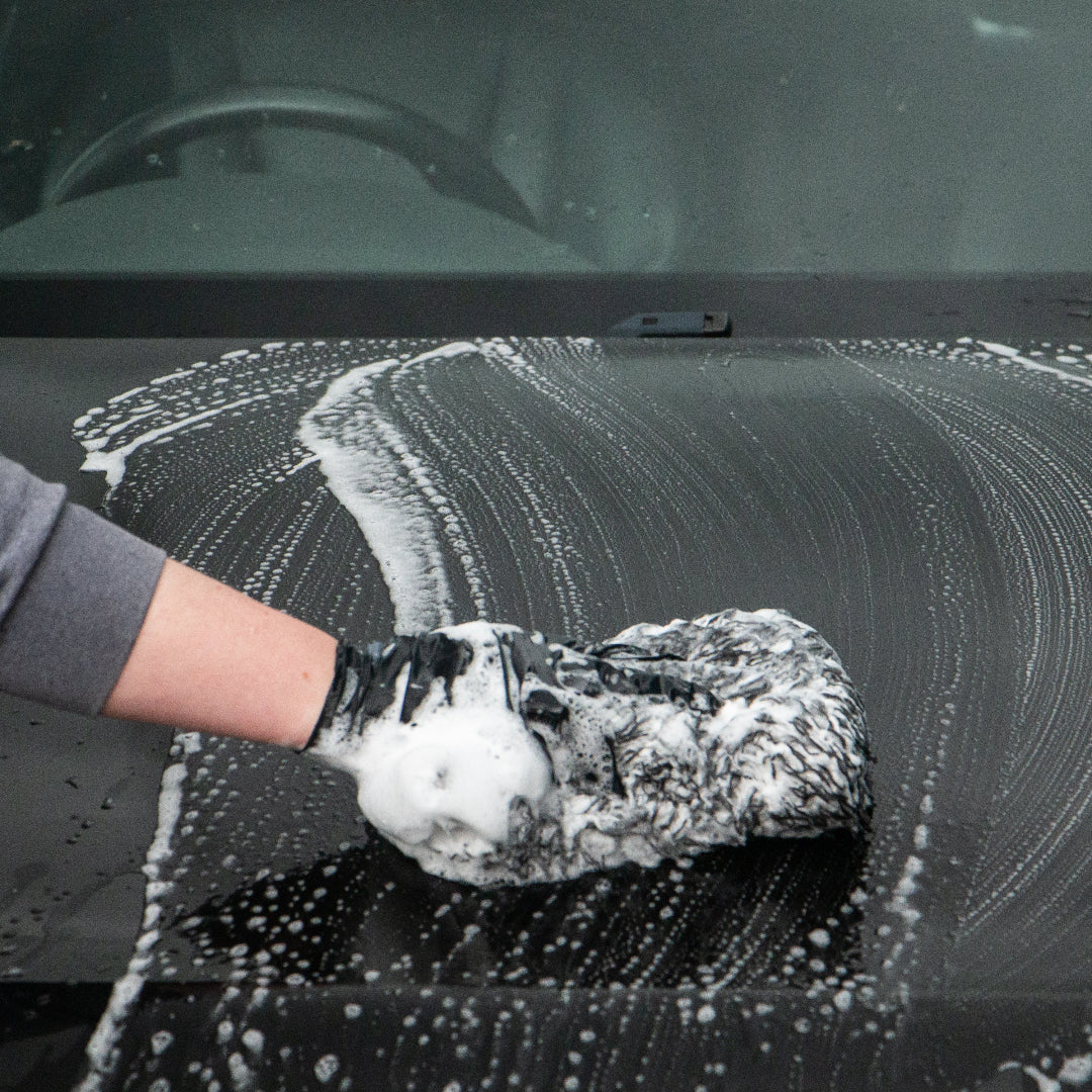A person wearing a grey long-sleeve shirt and a black glove washes a black car bonnet with soapy white foam using a textured sponge.