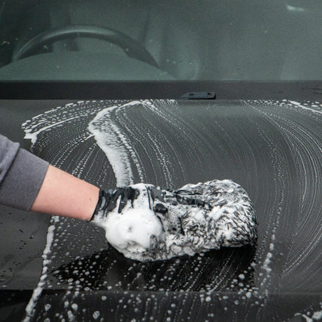 A person wearing a grey long-sleeve shirt and a black glove washes a black car bonnet with soapy white foam using a textured sponge.