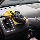 A person wearing a black glove wipes a grey car dashboard with a yellow microfiber cloth near the air vent.