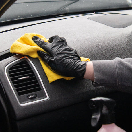 A person wearing a black glove wipes a gray car dashboard with a yellow microfiber cloth near the air vent.