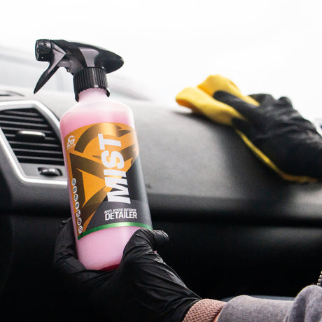 A person wearing black gloves holds a bottle of MIST Car Detailer with pink liquid inside a car, while wiping a black dashboard with a yellow cloth.