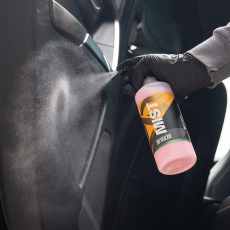 A person wearing a black glove sprays interior cleaner  labeled MIST onto a dark car door panel.
